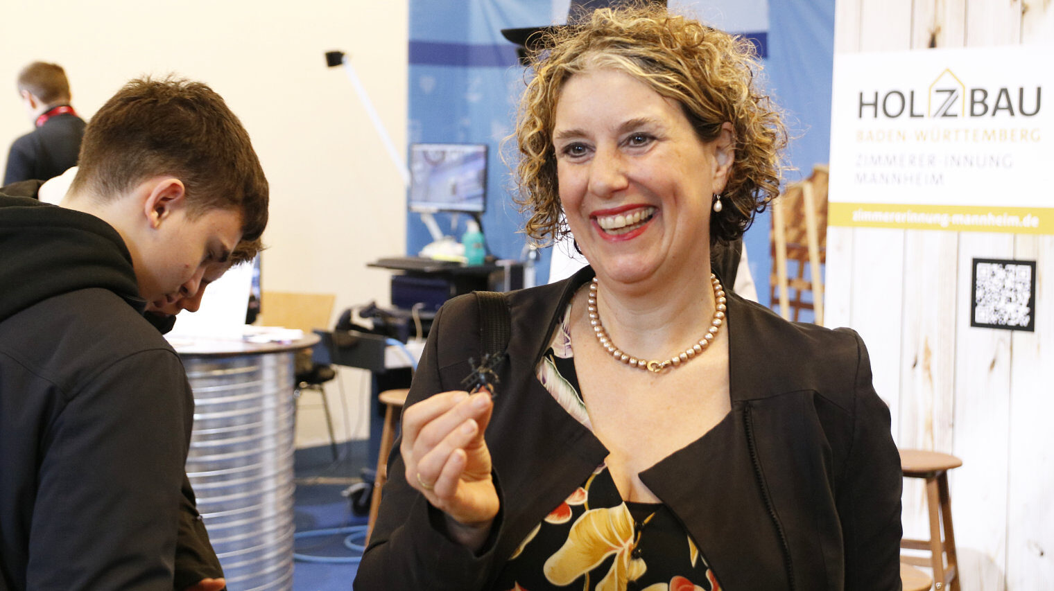 Frau mit gelocktem, kurzem Haar und einem strahlenden Lachen auf einer Messe, hält eine winzige Schornsteinfeger-Figur in der Hand und zeigt diese, links davon schauen Jugendliche auf den Stand