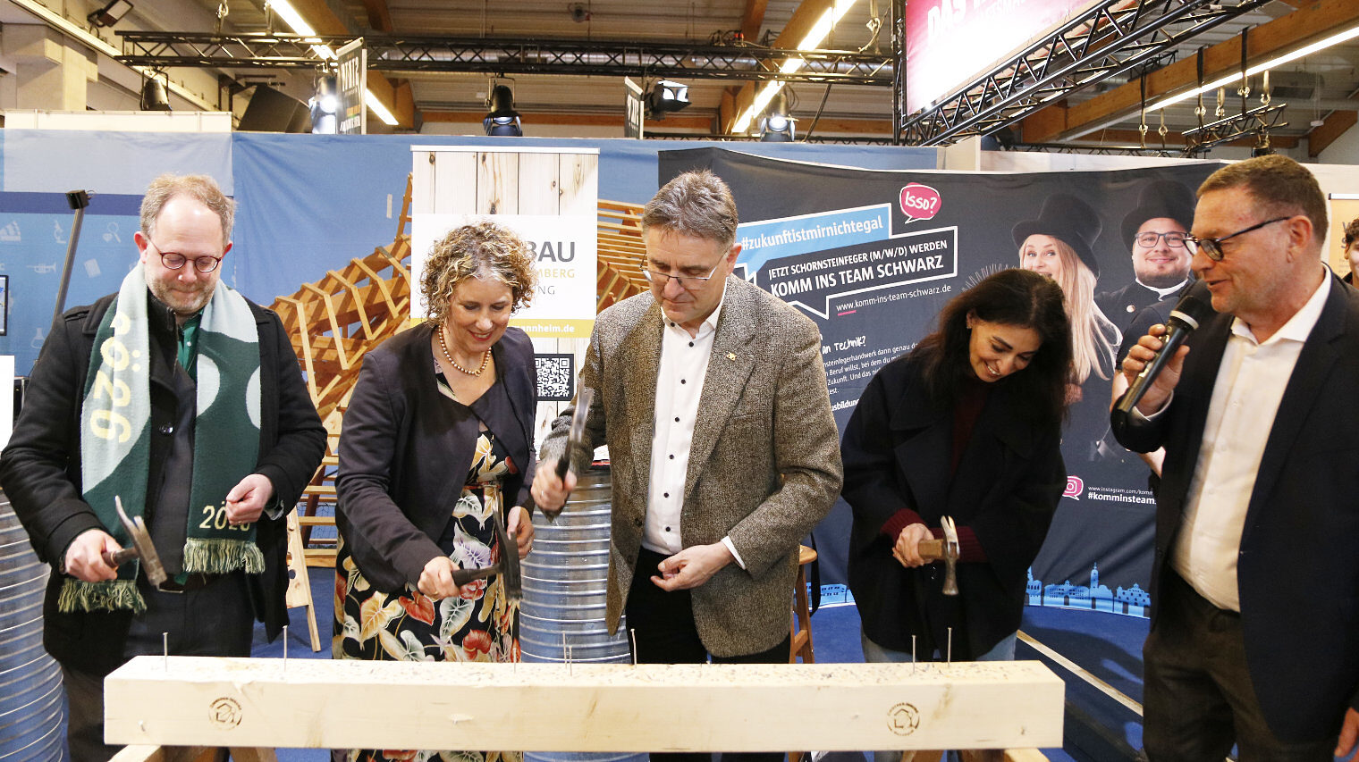 4 Personen stehen vor einem Holzbalken auf einer Messe und schlagen mit dem Hammer Nägel ein, rechts steht ein Mann mit Brille und moderiert
