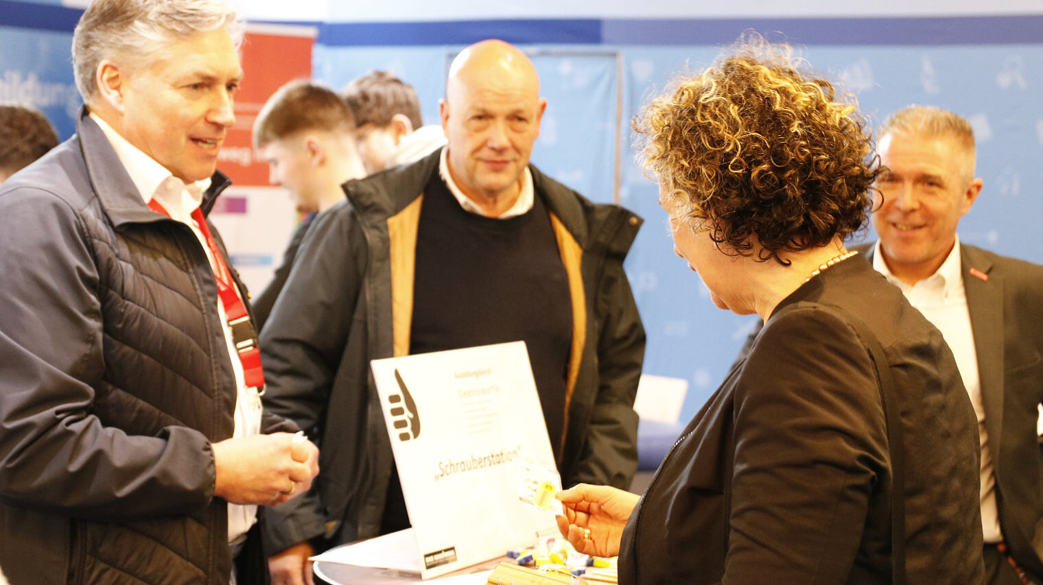 Messeszene an einem Stand, links zwei Männer, einer erklärt etwas, vorne mit dem Rücken zur Kamera eine Frau mit lockigen Haaren, die etwas betrachtet, rechts im Hintergrund ein weiterer Mann