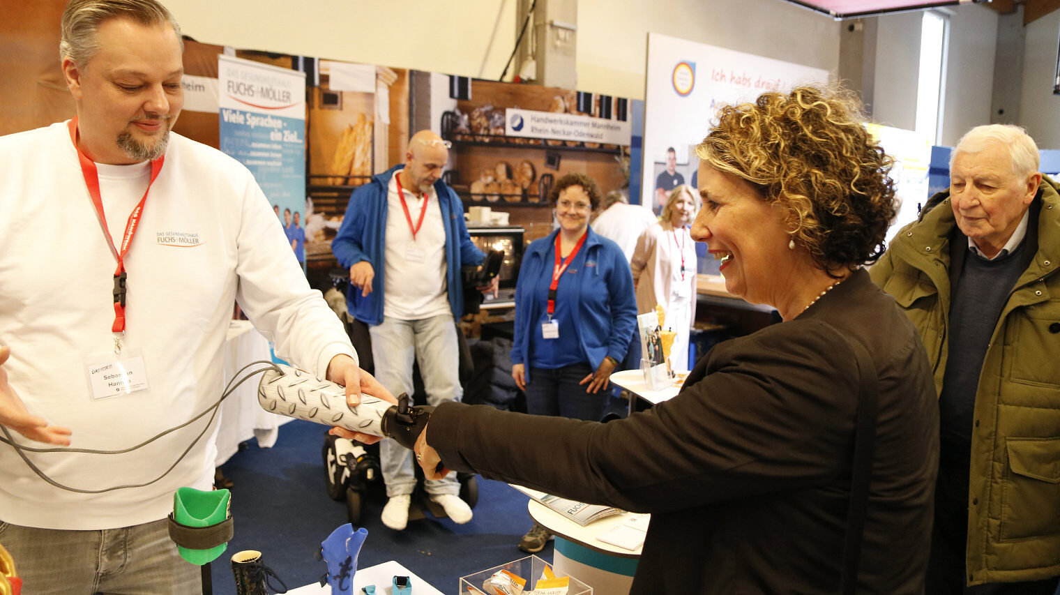 Messeszene an einem Stand, links ein Mann mit rotem Bändel um den Hals und einem bionischen Arm in der Hand, rechts eine Frau, die ihn lachend betrachtet, im Hintergrund weitere Menschen