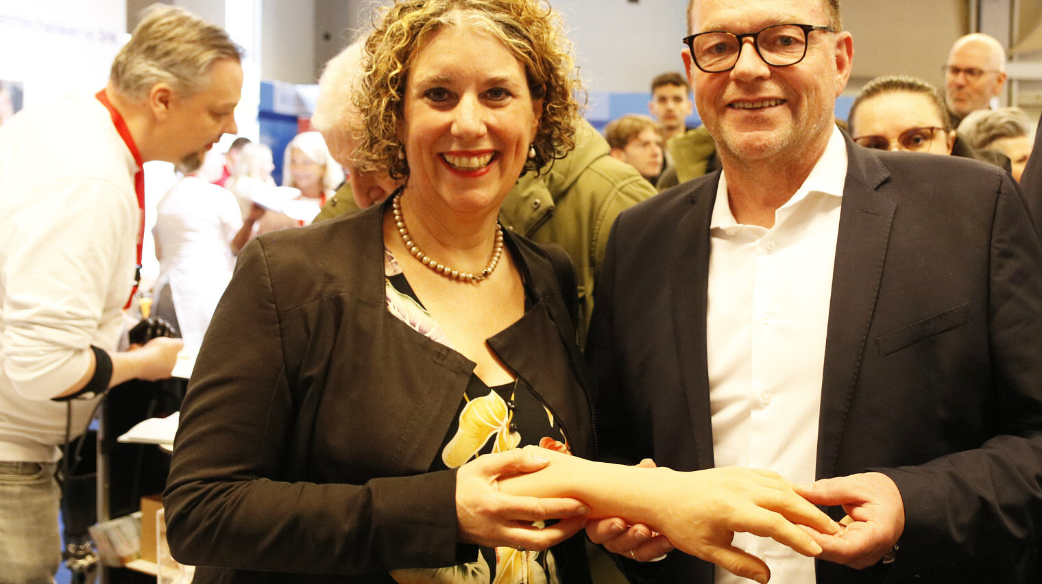 Eine Frau und ein Mann stehen lachend nebeneinander und schauen in die Kamera, zusammen halten sie eine orthopädische Hand, im Hintergrund eine Messeszene