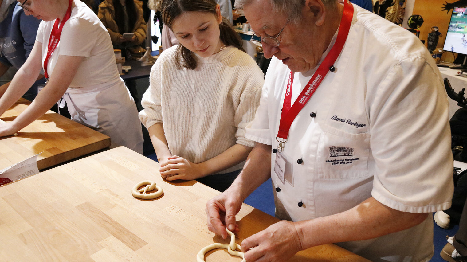 Bäcker an einem Messestand legt Teig zu einer Brezel, links neben ihm ein Mädchen, das ihn gespannt beobachtet, im Hintergrund eine Bäckerin beim Teigrollen