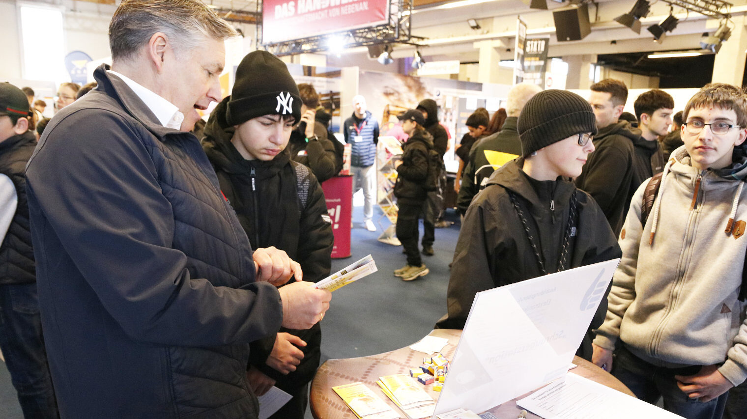 Messeszene mit Blick in die Halle und vielen jungen Leuten, links im Vordergrund ein älterer Mann, der einem Jugendlichen mit Mütze einen Flyer zeigt, andere Jugendliche stehen mit Abstand dabei
