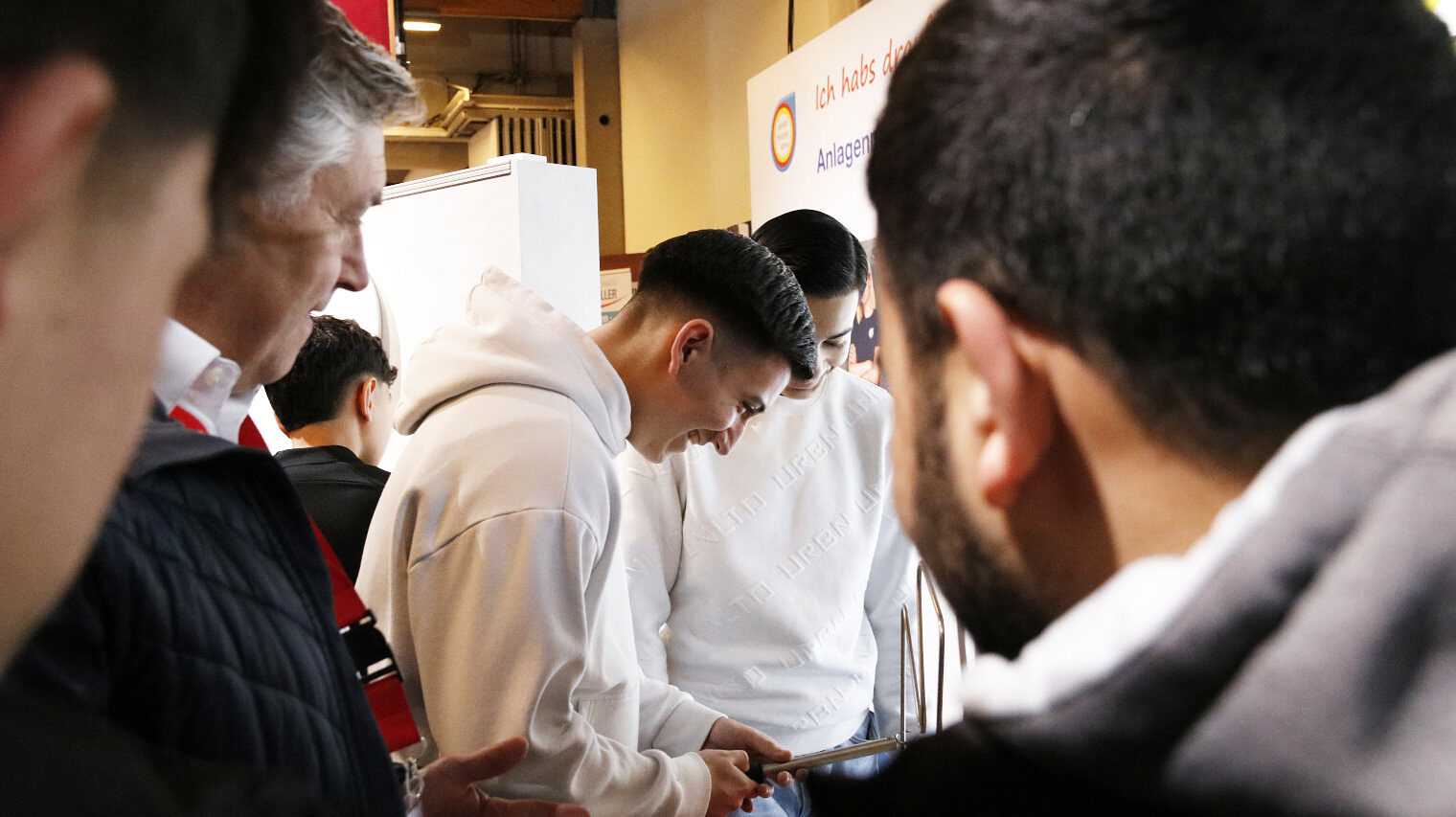 Blick über die Schulter von Männern im Vordergrund auf einen Messestand, an dem zwei junge Männer mit einem Stab entlang einer Spirale fahren und dabei lachen