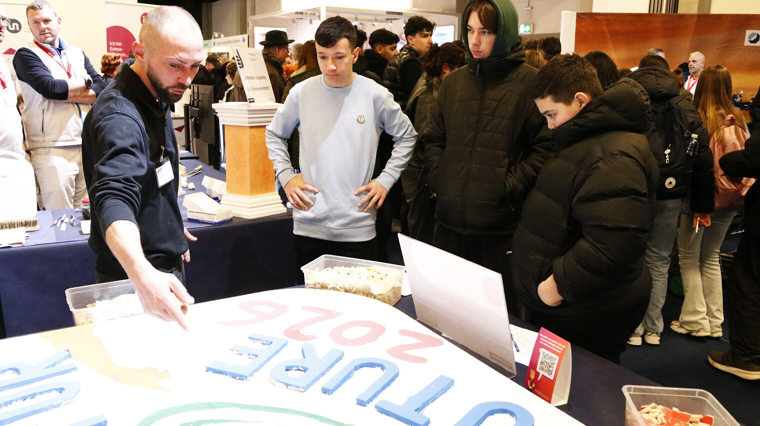 Messeszene mit Blick auf einen Stand, auf einer Tafel im Vordergrund bunte Buchstaben, ein Mann mit Glatze links zeigt etwas, drei Jungs in dicken Jacken schauen auf die Platte