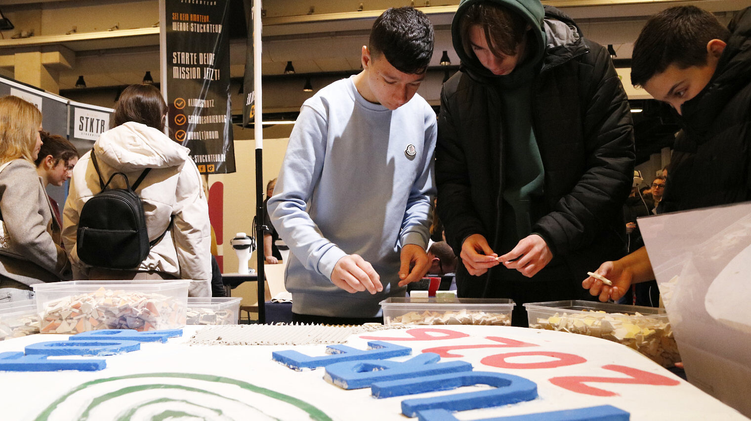Platte mit bunten Buchstaben im Vordergrund, daran stehen drei Jungs in dicken Jacken, die Fliesenteile in ihren Händen halten und schauen, wo sie diese hinlegen können, im Hintergrund andere