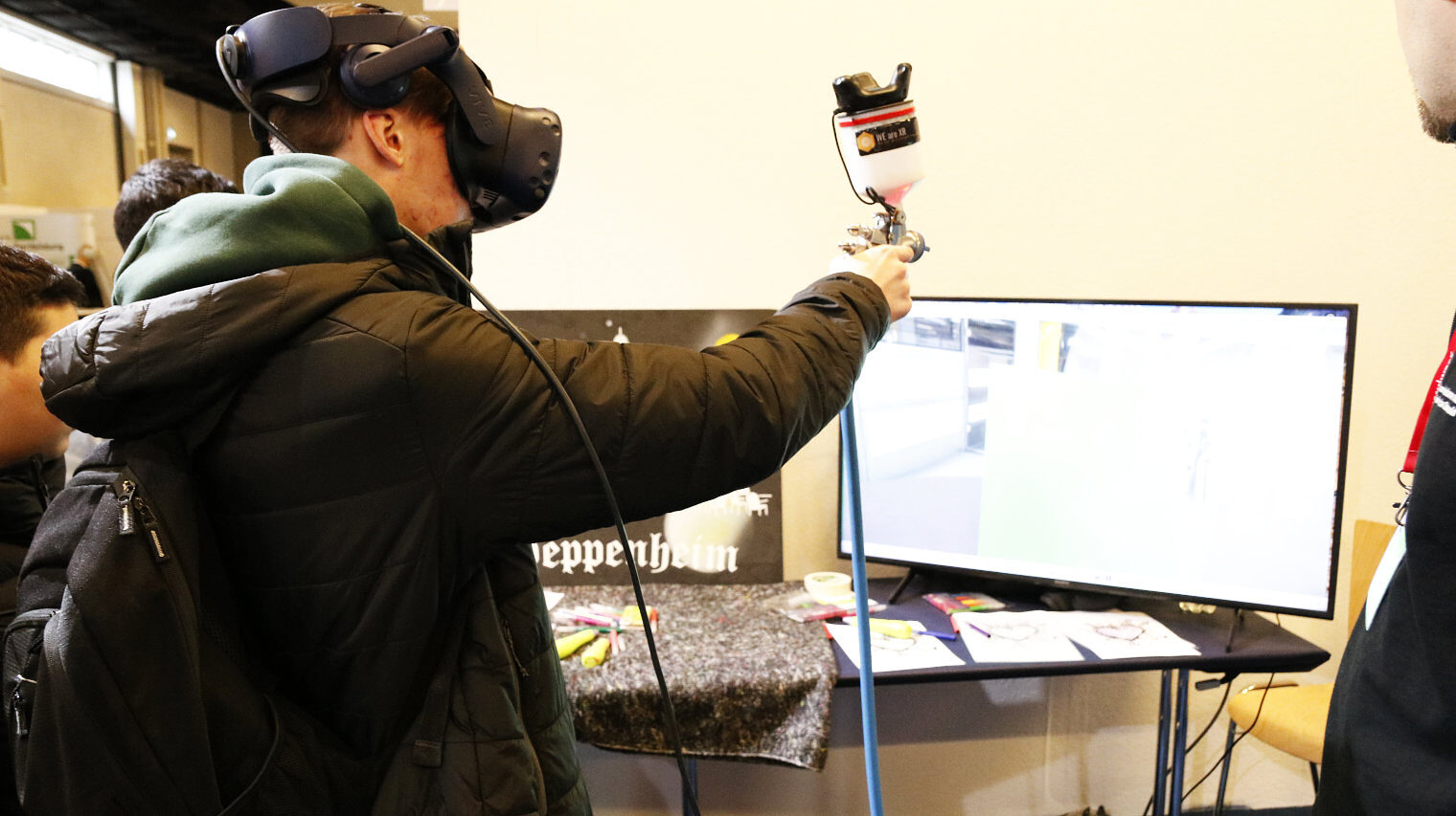 Junger Mann in dicker Winterjacke mit Kapuze auf einem Messestand, VR-Brille auf und eine Sprühpistole in der ausgestreckten Hand, vor ihm ein Bildschirm, recht ein Mann im Anschnitt