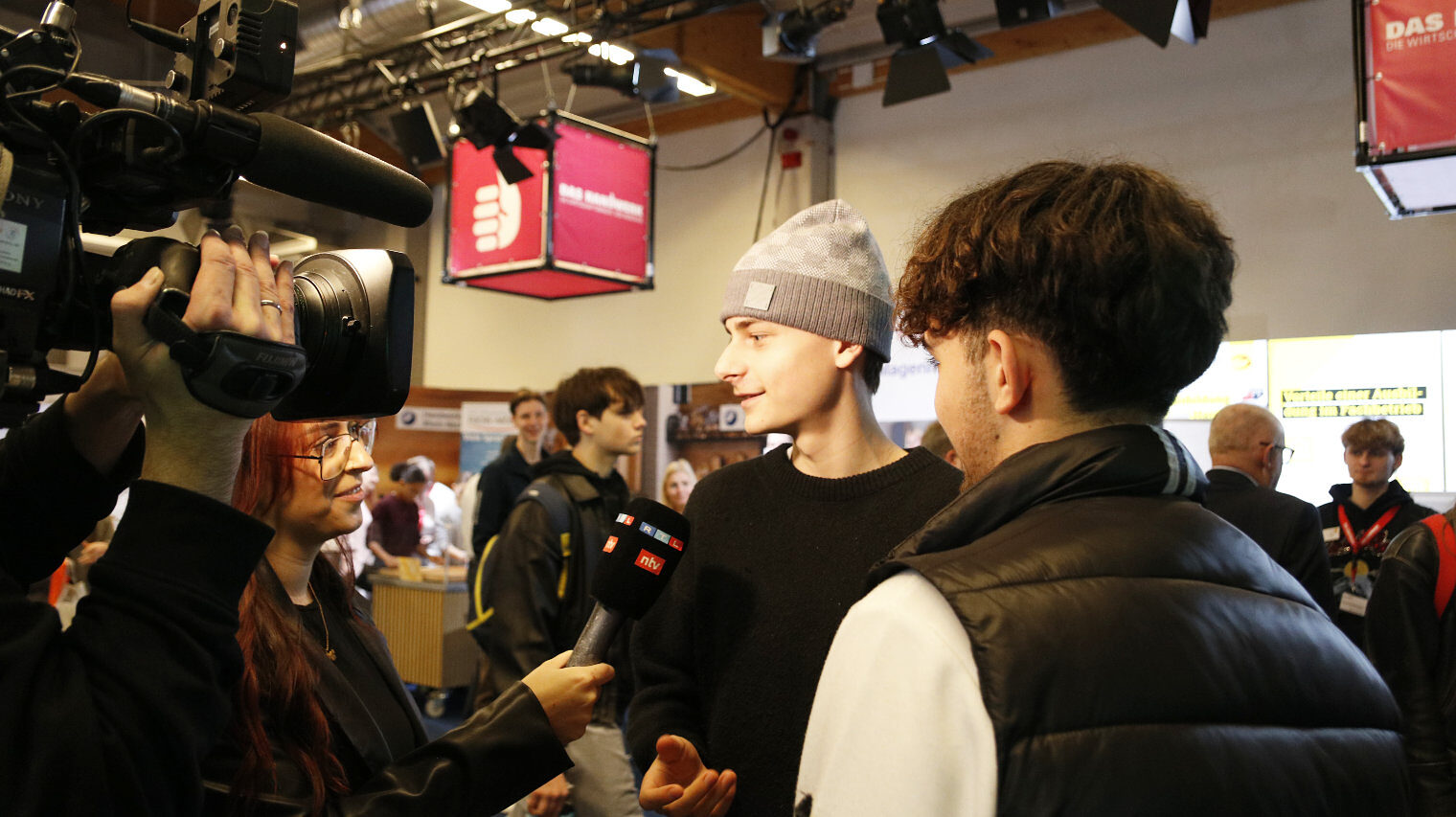 Reporterin mit Mikro in der Hand interviewt 2 junge Männer, einer mit Mütze, der rechts mit dem Rücken im Bild, links ragt eine Kamera ins Bild, die jemand auf der Schulter trägt