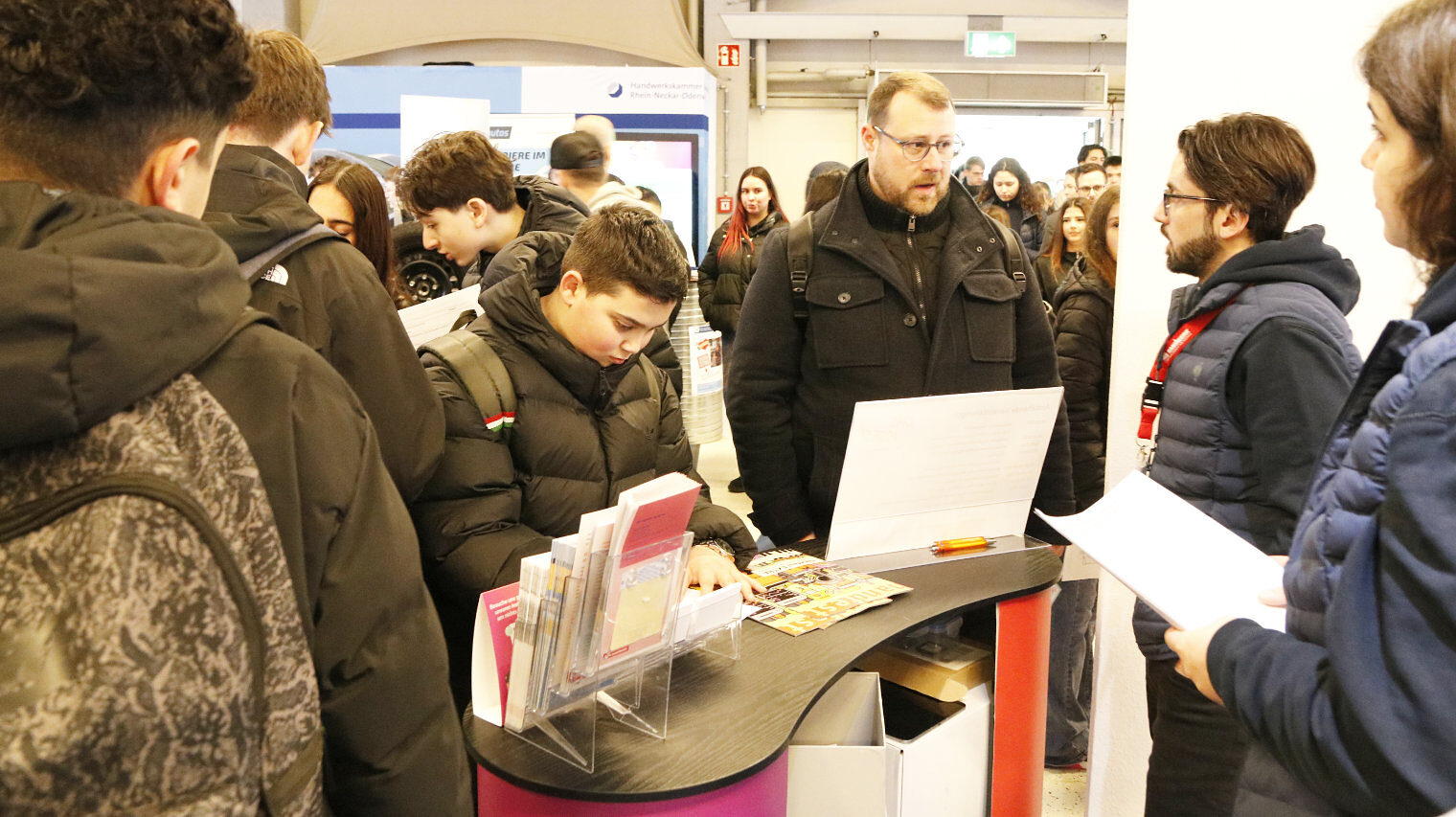 Messeszene mit einem Stand und regem Betrieb, zwei Menschen am Stand im Gespräch mit den Leuten vor ihnen, viele junge Leute rundherum, Trubel