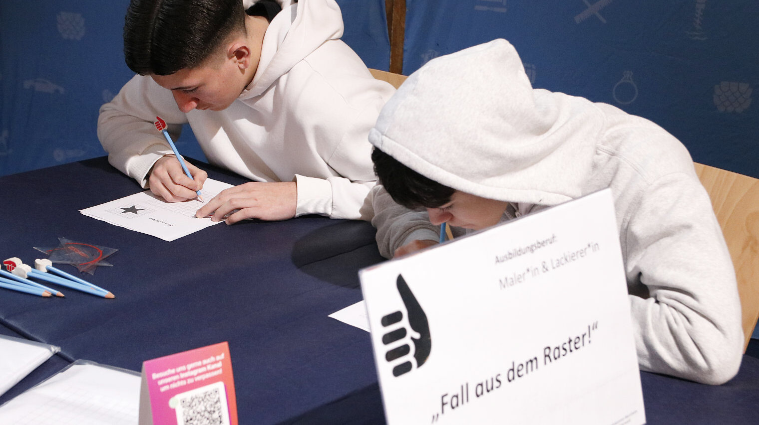 Zwei Jungs sitzen an einem Tisch, einer hat eine Kapuze über den Kopf gezogen, beide schreiben auf einem Papier vor sich, im Vordergrund ein Schild mit der Aufschrift Fall aus dem Raster