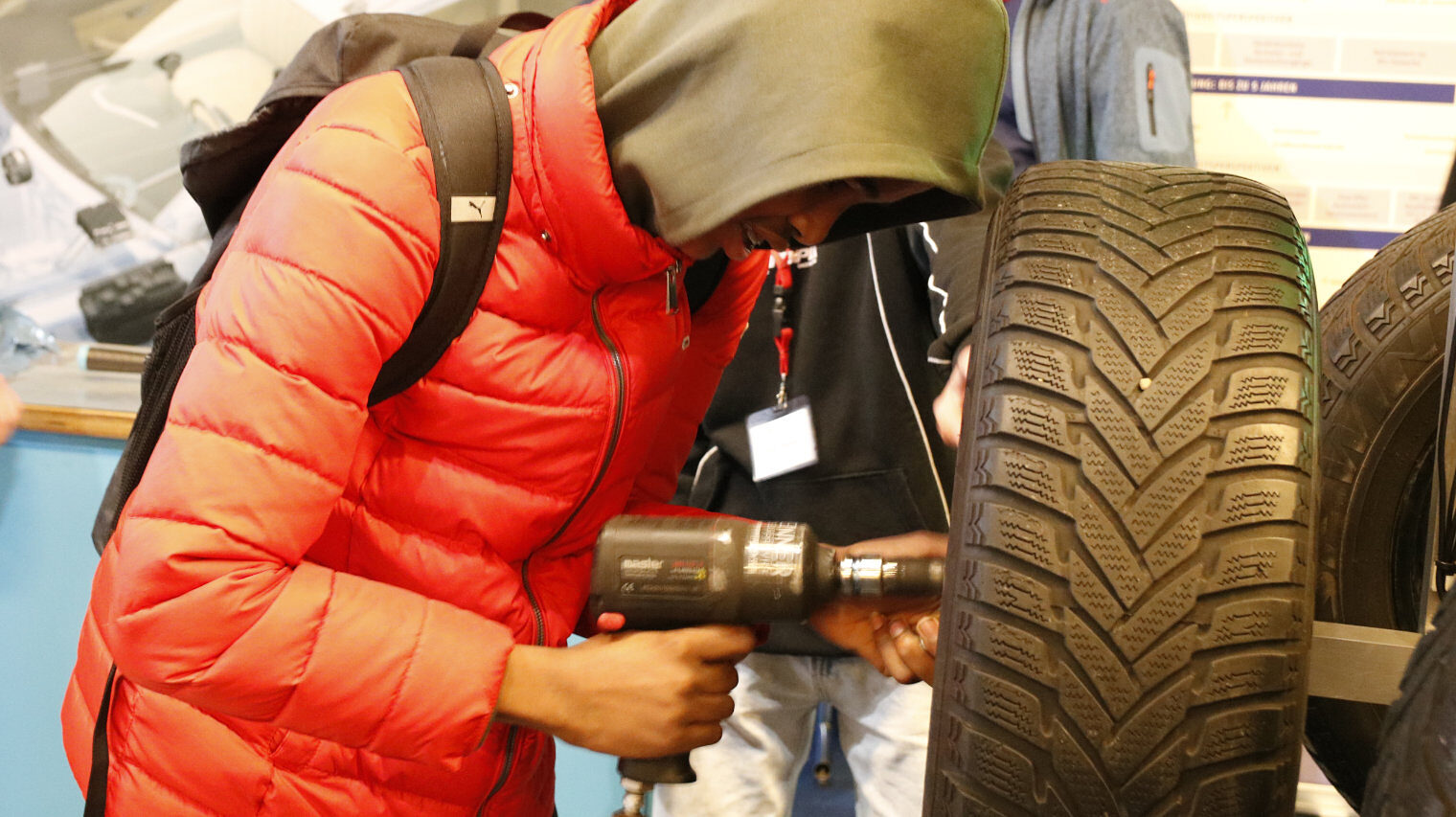 Jugendlicher in orangener Winnterjacke mit Kapuzenpulli über dem Kopf vor einer Autoreifen, in der Hand den Schrauber, Gesicht kaum zu sehen, arbeiten konzentriert am Reifen