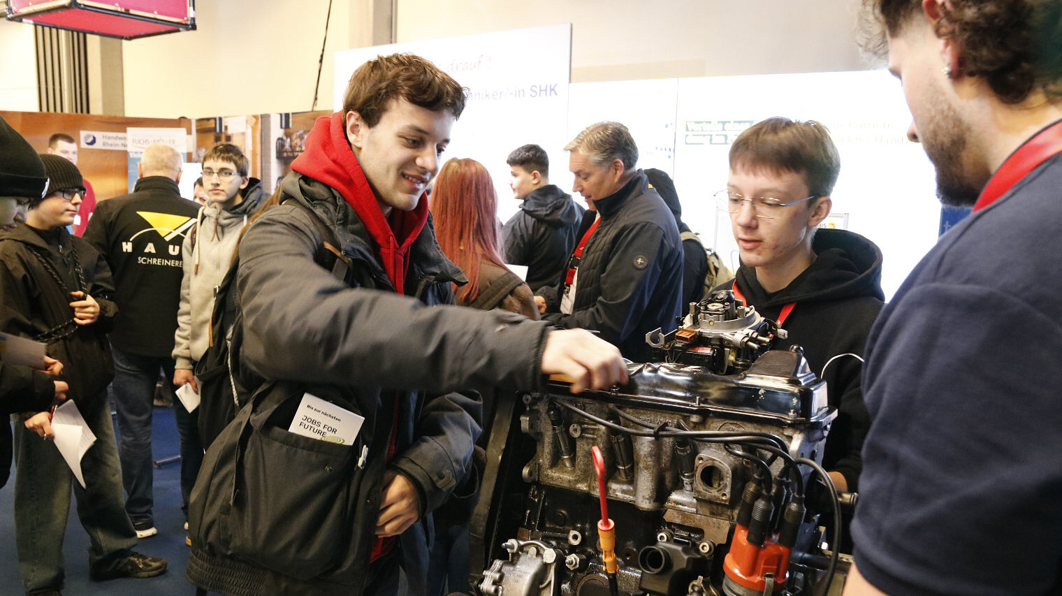 Junger Mann greift auf einem Messestand über einen Motor, dahinter ein Jugendlicher, der gespannt zusieht, rechts ein erwachsener Mann im Bildanschnitt, im Hintergrund Messetrubel