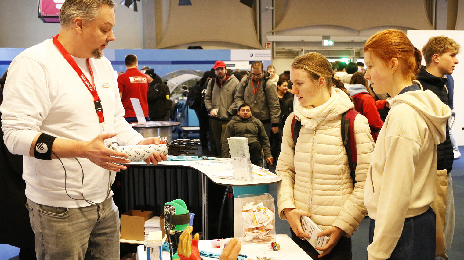 Links ein Mann mit grauem Kinnbart, weißem Pulli, rotem Messeband um den Hals, verkabelt am Arm, zeigt zwei Mädchen vor dem Messestand eine Prothese