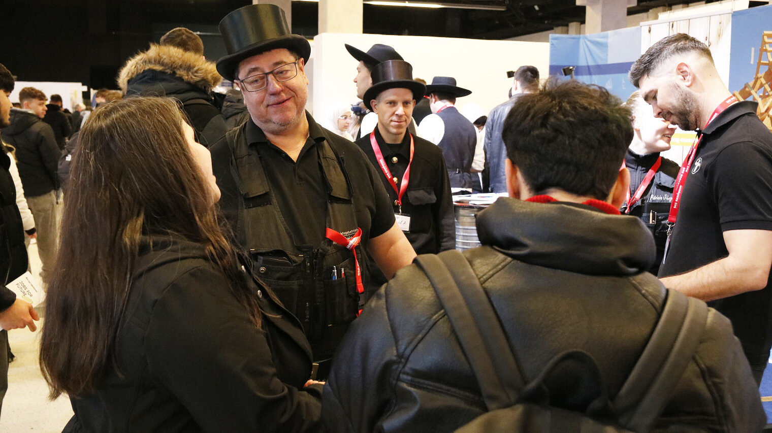 Messeszene am Stand der Schornsteinfeger, 2 Schornsteinfeger in der typischen Kluft sprechen mit zwei jungen Leuten, die mit dem Rücken zur Kamera stehen, Hintergrund Messeambiente