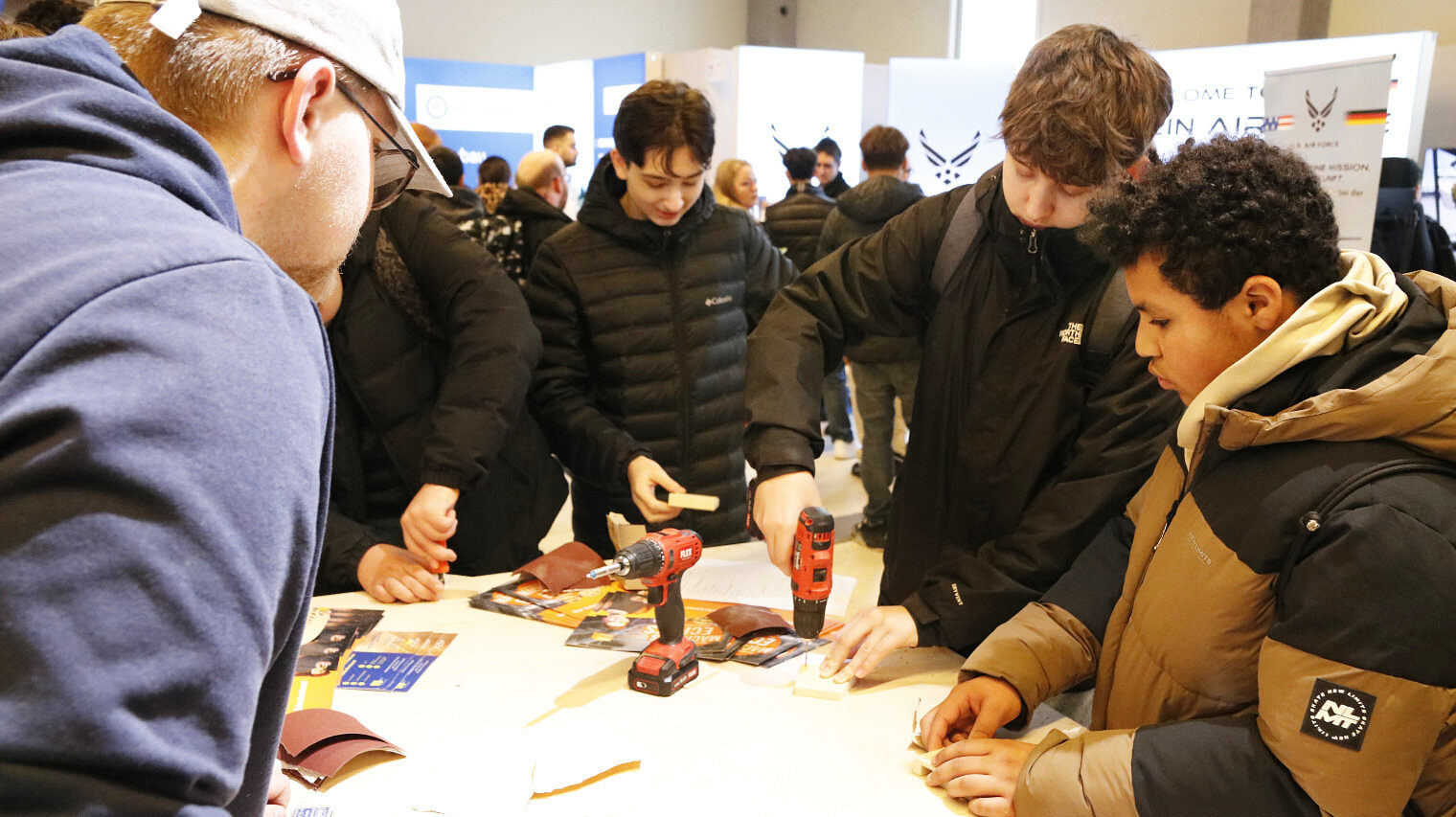 belebte Messeszene, links angeschnitten ein Mann mit Cap im blauen Kapuzenpulli, beugt sich über den Tisch zu Jungs auf der rechten Seite, einer mit kleinem Bohrer in der Hand bei der Arbeit