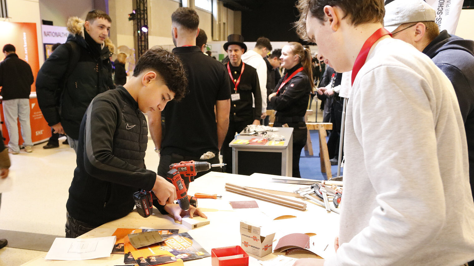 Messeszene, Stand mit kleinen Holzbauteilen, rechts Messepersonal, links ein Junge mit Bohrer in der Hand bei der Arbeit, Hintergrund Personen aus der Messe