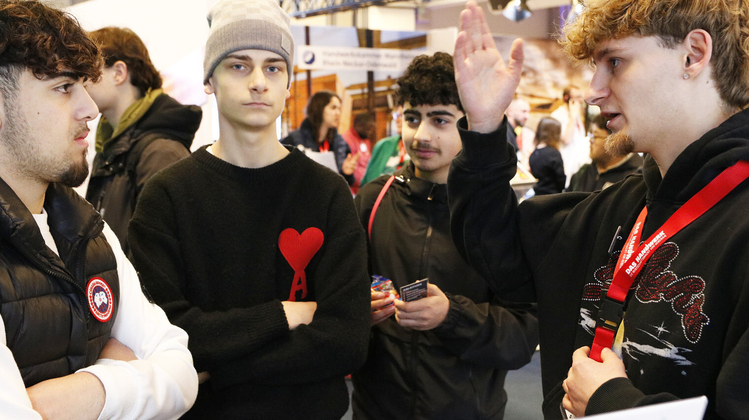 Gruppe jugendlicher Männer auf einer Messe, rechte Person mit rotem Messehalsband, mit einer Hand zeigt er etwas und spricht, Person gegenüber mit verschränkten Armen hört aufmerksam zu