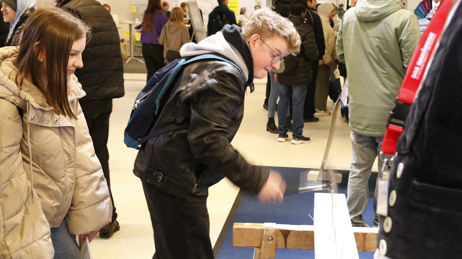 Messestand, Junge mit blondem Haar, Brille, Rucksack über der dunklen Jacke schlägt mit dem Hammer einen Nagel in den Balken auf einem Bock, links schaut ein Mädchen lachend zu