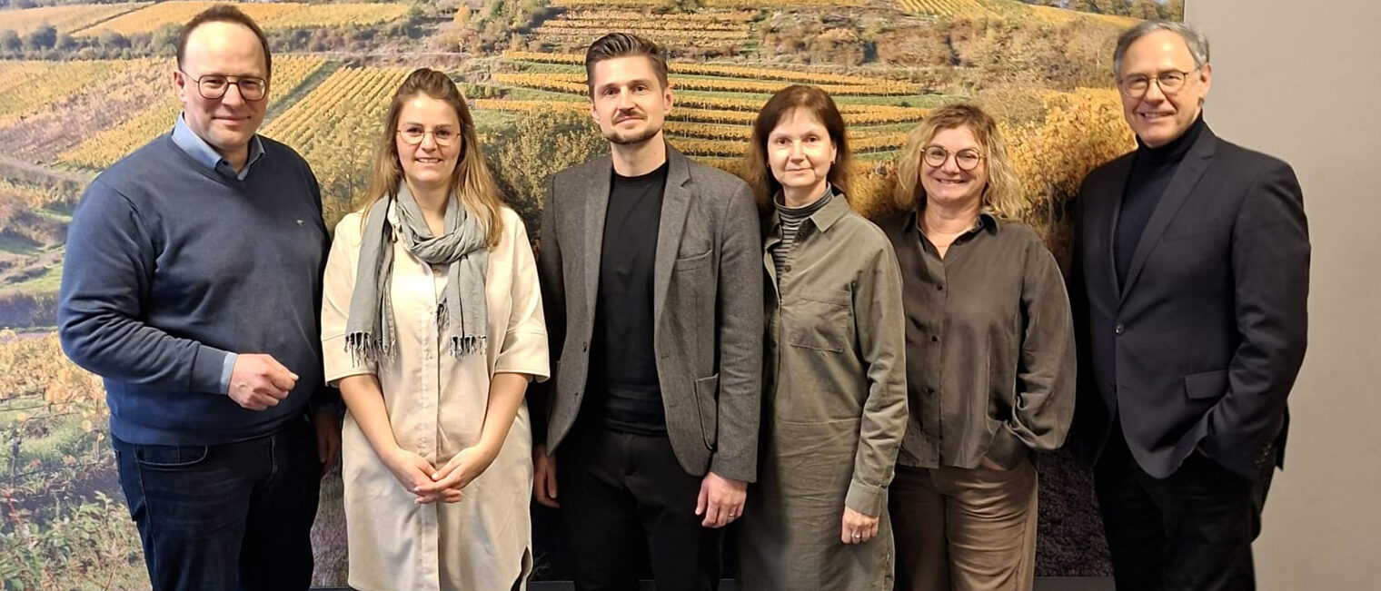 Sechs Personen vor einer Bilderwand mit Weinberg im Herbst, links, in der Mitte und rechts Männer, dazwischen Frauen, alle schauen lächelnd in die Kamera