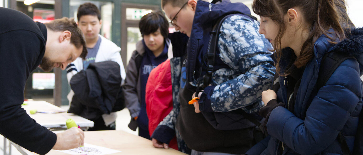 Gruppe von Kindern in Winterjacken steht rechterhand an einem Werkstatt-Tisch über den sich von links ein Mann mit Bart beugt und mit einem Stift in der Hand über ein Blatt Papier fährt.