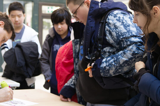 Gruppe von Kindern in Winterjacken steht rechterhand an einem Werkstatt-Tisch über den sich von links ein Mann mit Bart beugt und mit einem Stift in der Hand über ein Blatt Papier fährt.