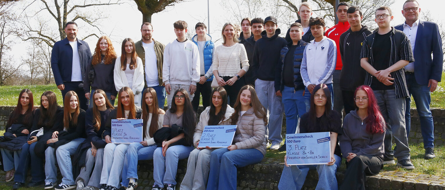 27-köpfige Gruppe mit Schülerinnen, Schülern und Lehrern im Freien, vordere Reihe sitzt auf einer Stufe, drei große Schecks, hintere Reihe stehend, alle schauen in die Kamera