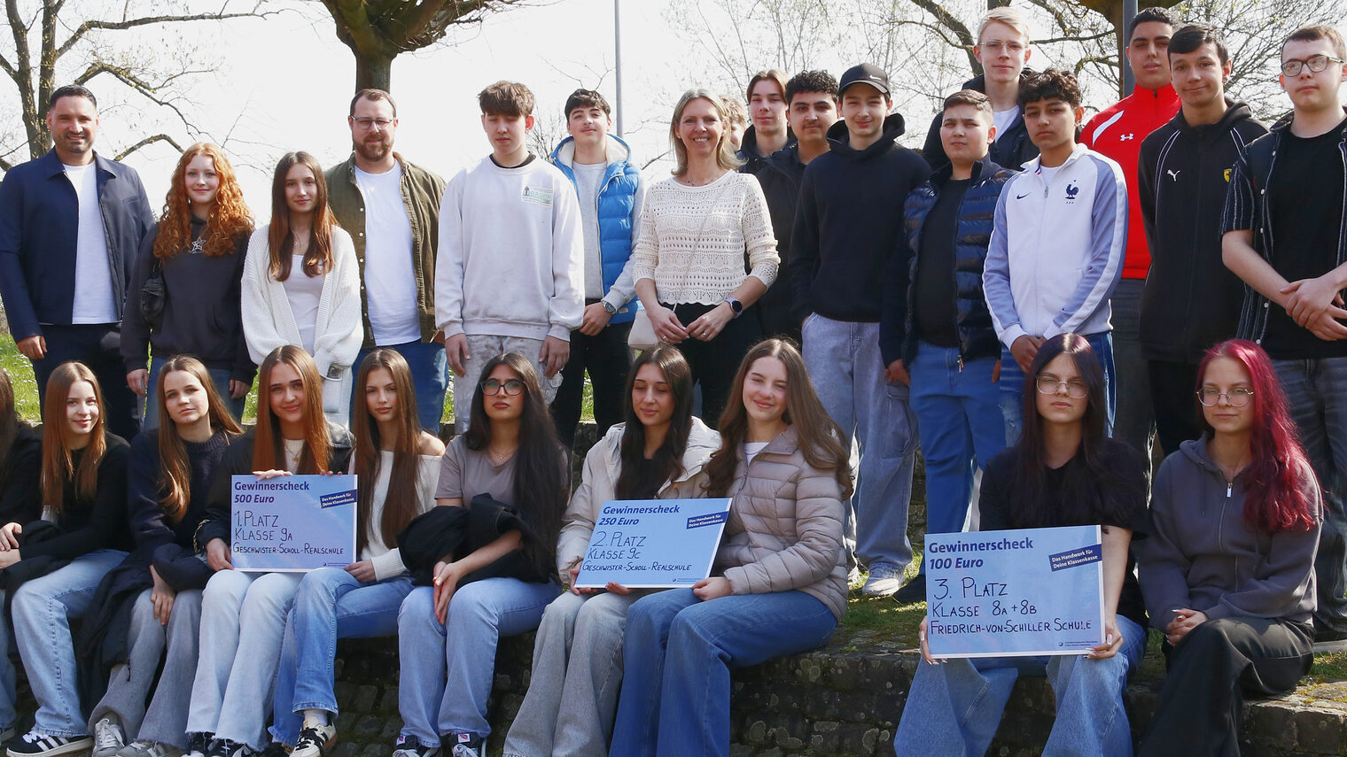 27-köpfige Gruppe mit Schülerinnen, Schülern und Lehrern im Freien, vordere Reihe sitzt auf einer Stufe, drei große Schecks, hintere Reihe stehend, alle schauen in die Kamera