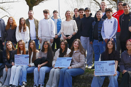 27-köpfige Gruppe mit Schülerinnen, Schülern und Lehrern im Freien, vordere Reihe sitzt auf einer Stufe, drei große Schecks, hintere Reihe stehend, alle schauen in die Kamera