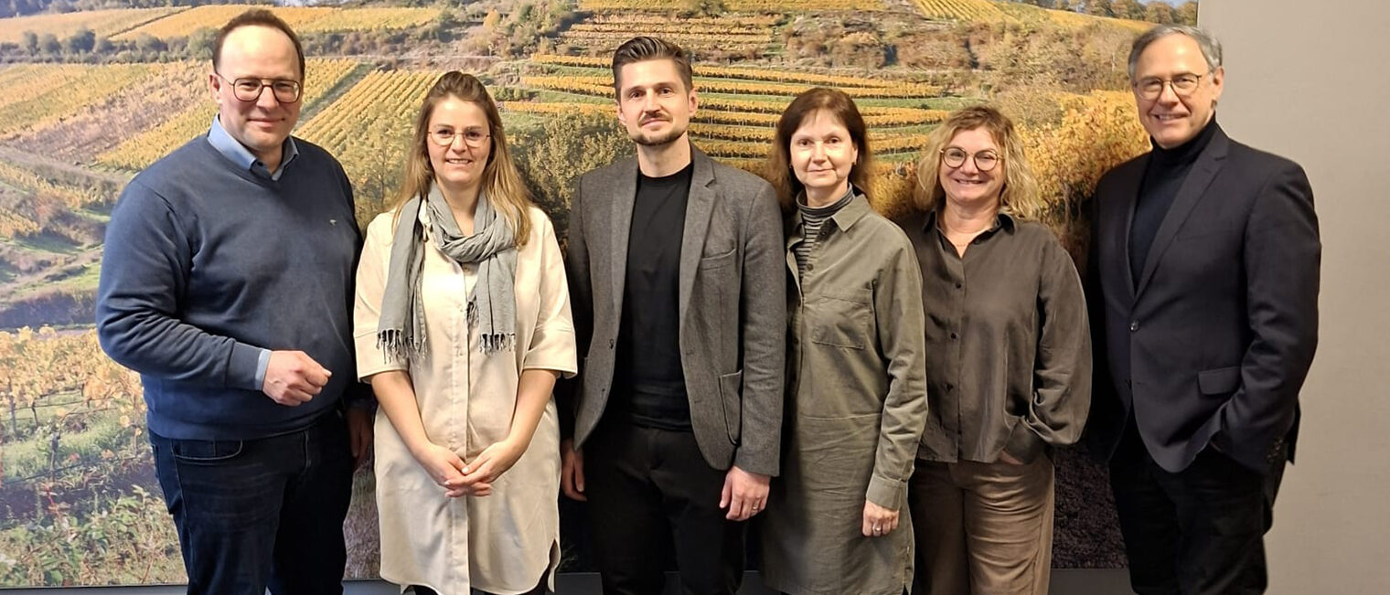 Gruppe aus 6 Personen vor einer Fotowand mit herbstlichem Weinberg-Motiv, außenstehend und in der Mitte Männer, dazwischen Frauen, alle schauen in die Kamera