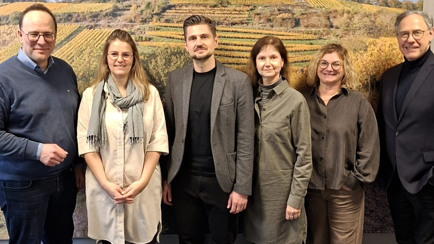Gruppe aus 6 Personen vor einer Fotowand mit herbstlichem Weinberg-Motiv, außenstehend und in der Mitte Männer, dazwischen Frauen, alle schauen in die Kamera