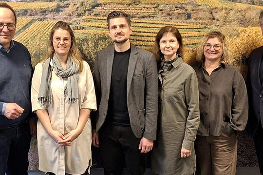 Gruppe aus 6 Personen vor einer Fotowand mit herbstlichem Weinberg-Motiv, außenstehend und in der Mitte Männer, dazwischen Frauen, alle schauen in die Kamera