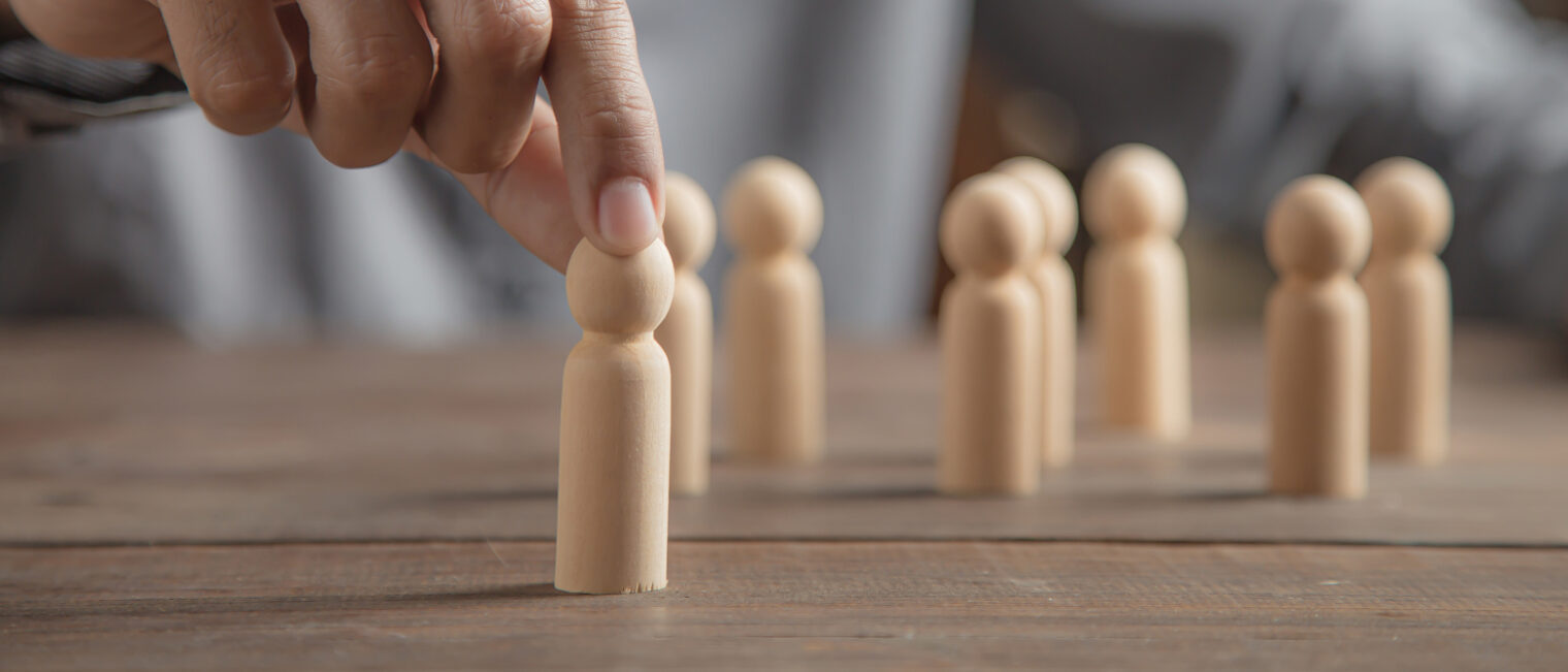 Hand die eine Holzfigur von einem Tisch anhebt, auf dem weitere Holzfiguren stehen.
