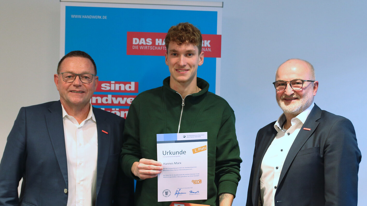 Preisübergabe, drei Männer stehen nebeneinander, der Herr in der Mitte hält sein Zeugnis und seinen Gutschein in die Kamera