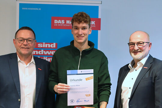 Preisübergabe, drei Männer stehen nebeneinander, der Herr in der Mitte hält sein Zeugnis und seinen Gutschein in die Kamera