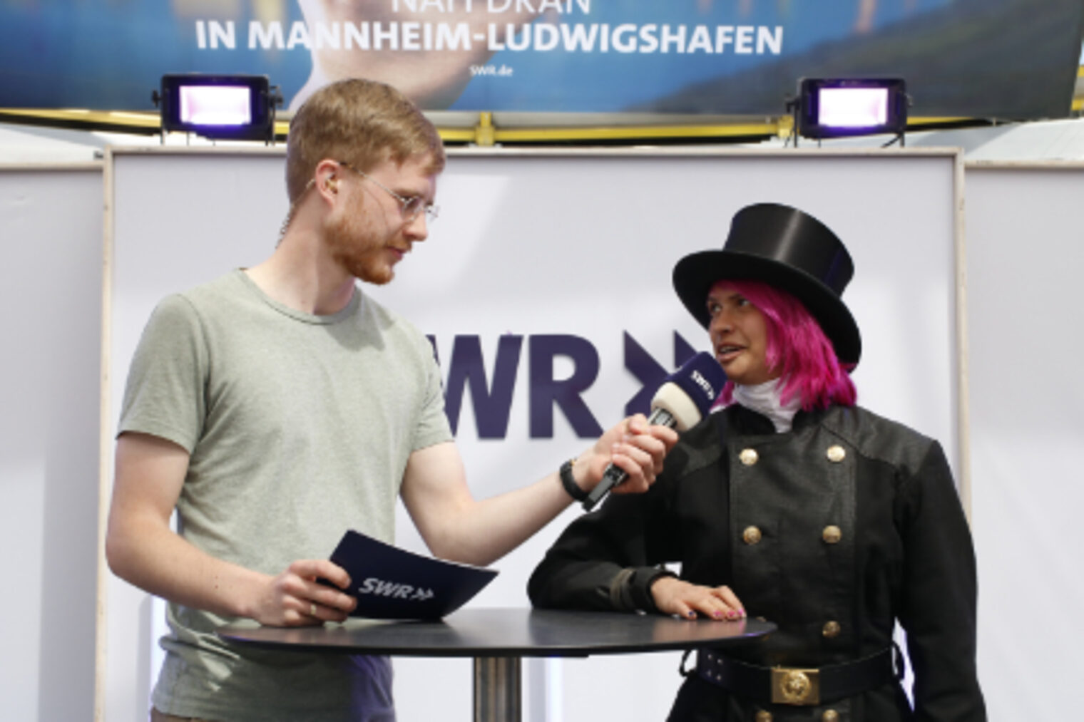 SWR-Moderator interviewt eine Schornsteinfegerin in Zunfttracht mit pinken Haaren am SWR-Stand auf dem Maimarkt Mannheim