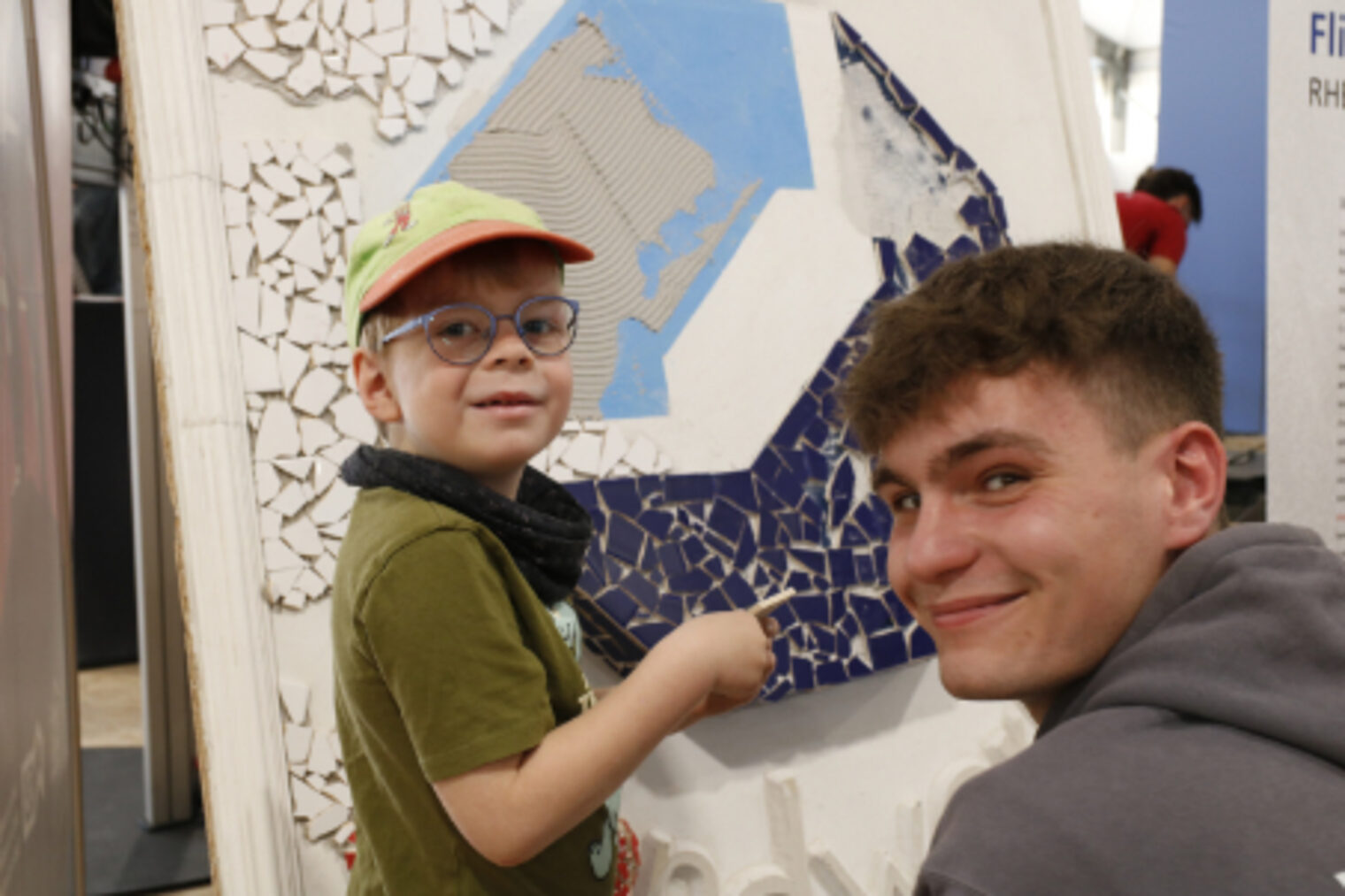 Kind und junger Mann zeigen gemeinsam lachend auf ein Mosaik-Kunstwerk am Fliesenleger-Stand auf dem Maimarkt Mannheim