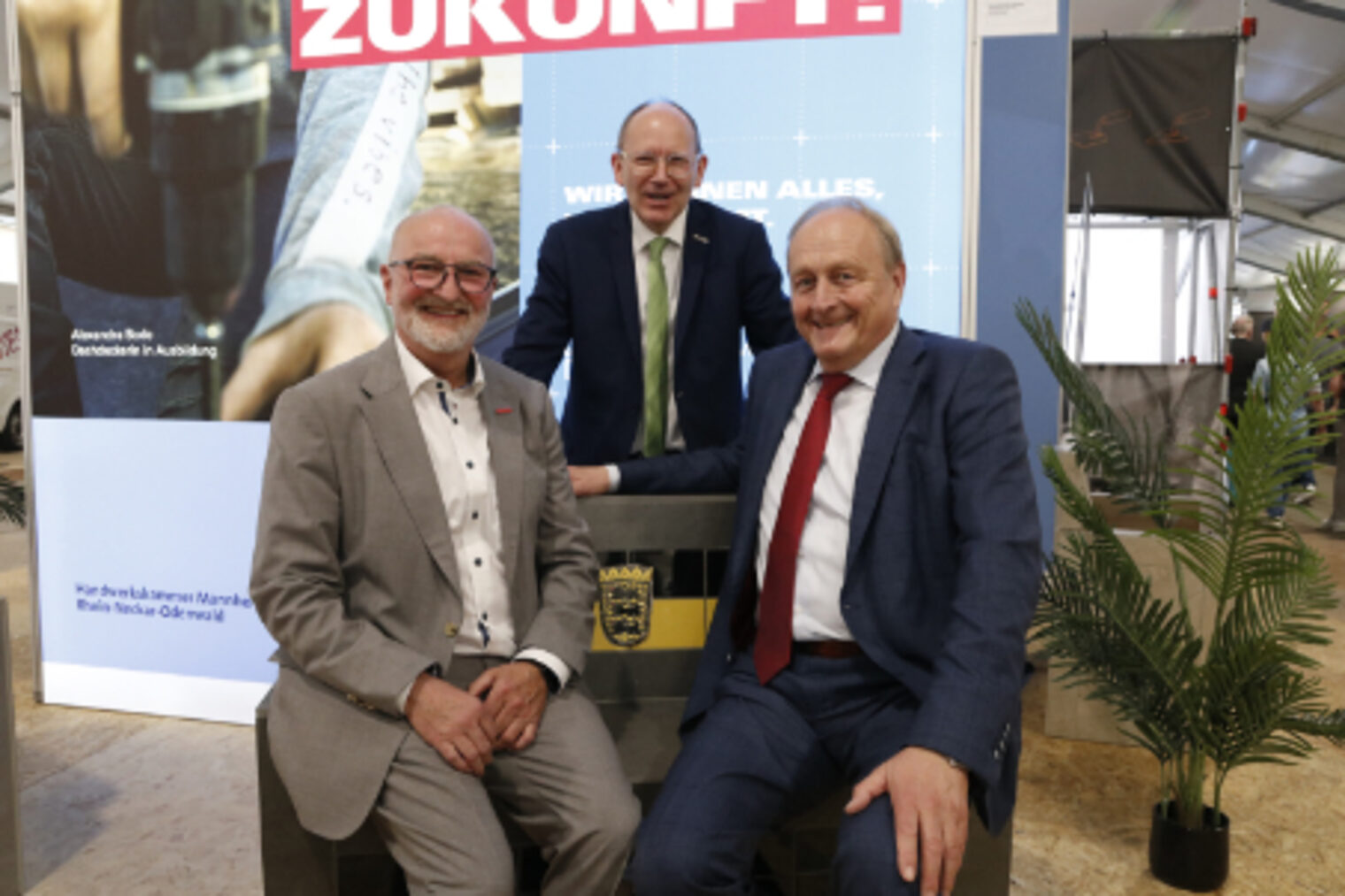 Drei Herren posieren laechelnd auf und neben einer gefliesten Sitzbank mit Baden-Wuerttemberg-Wappen am HWK-Stand auf dem Maimarkt Mannheim