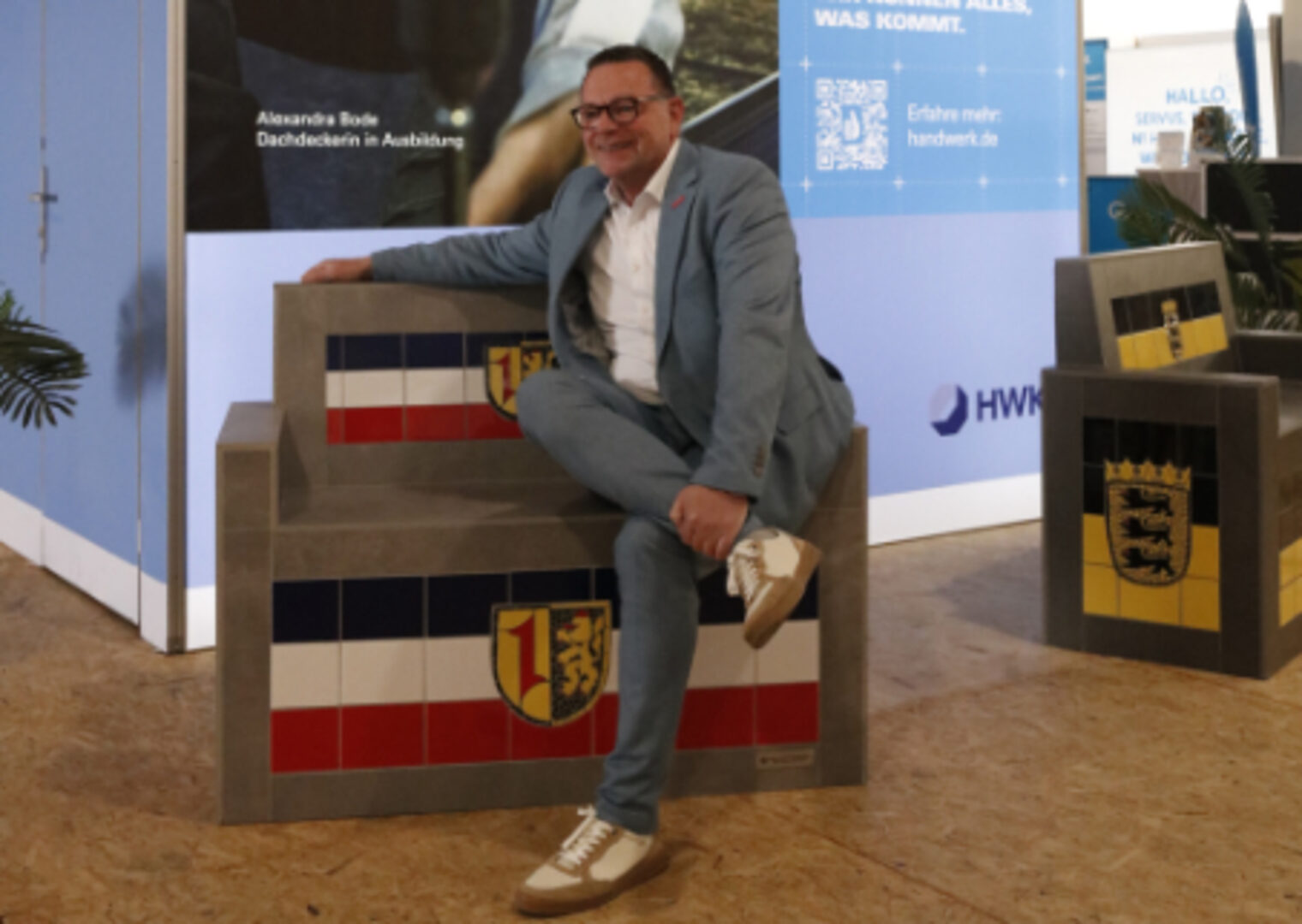 Präsident Klaus Hofmann sitzt laechelnd auf einer gefliesten Sitzbank mit Mannheimer Wappen am HWK-Stand auf dem Maimarkt Mannheim