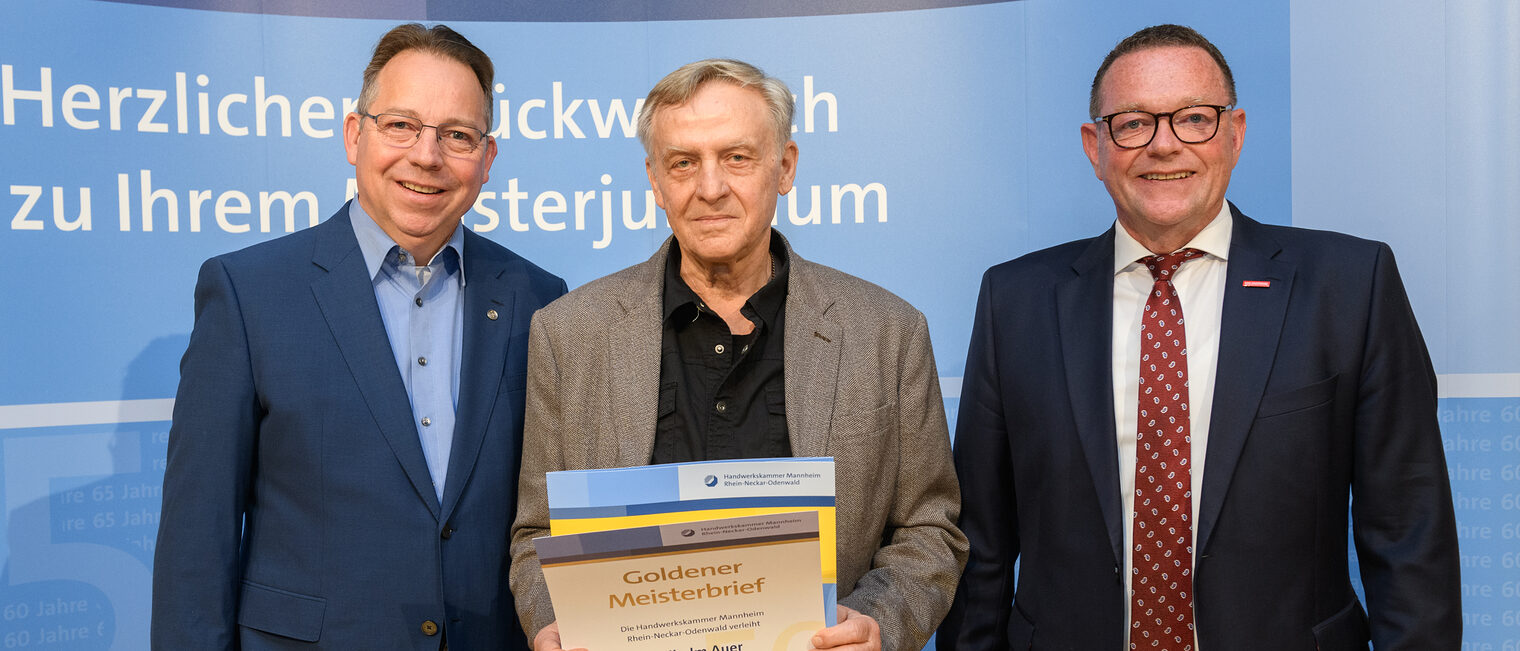 Drei Personen posieren für ein Foto, in der Mitter wird die Ehrenurkunde gehalten