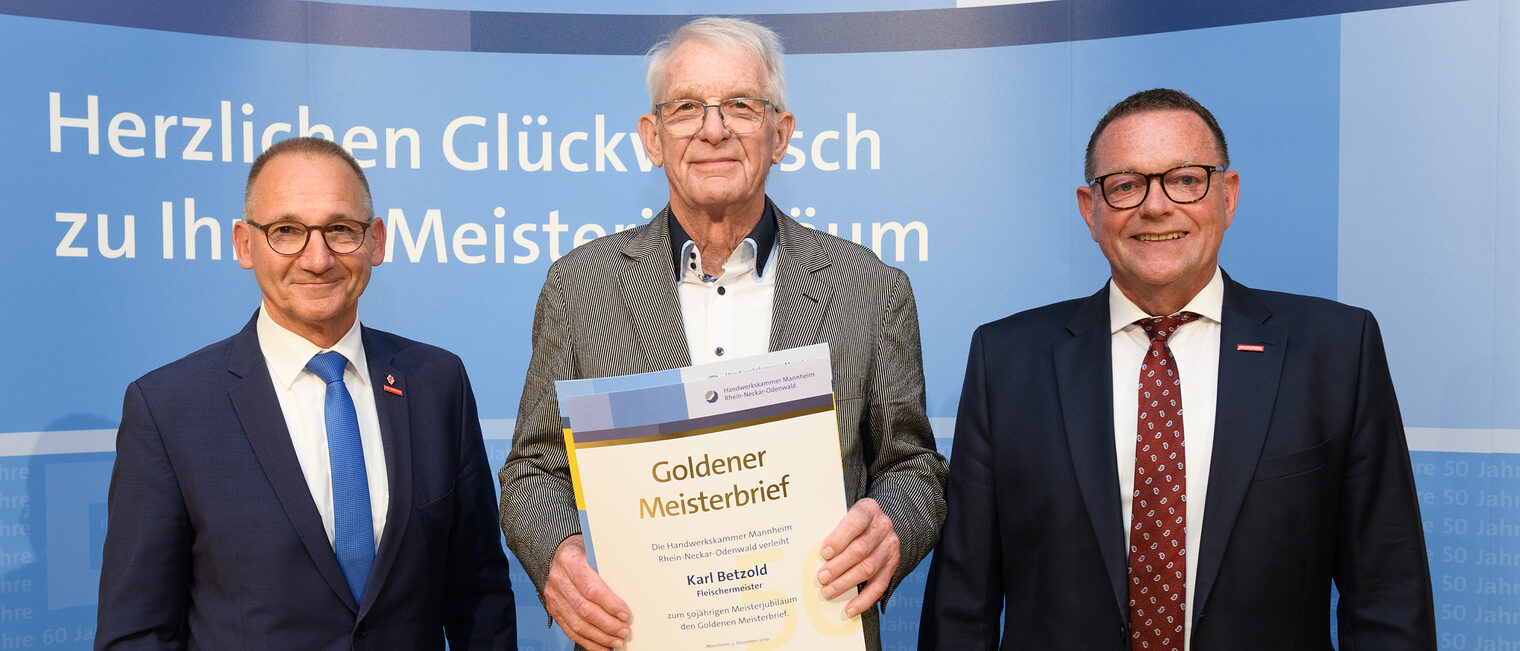 Drei Personen posieren für ein Foto, in der Mitte wird die Ehrenurkunde gehalten