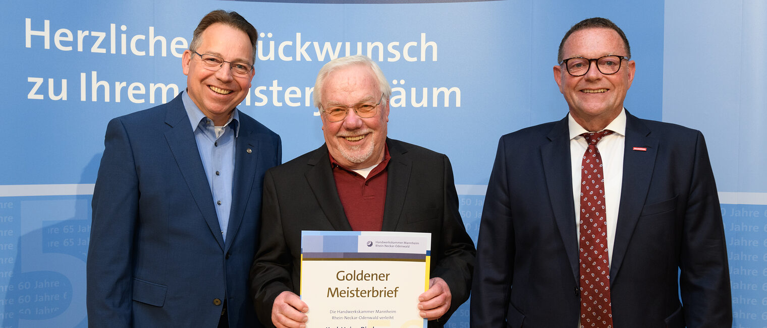 Drei Personen posieren für ein Foto, in der Mitte wird die Ehrenurkunde gehalten