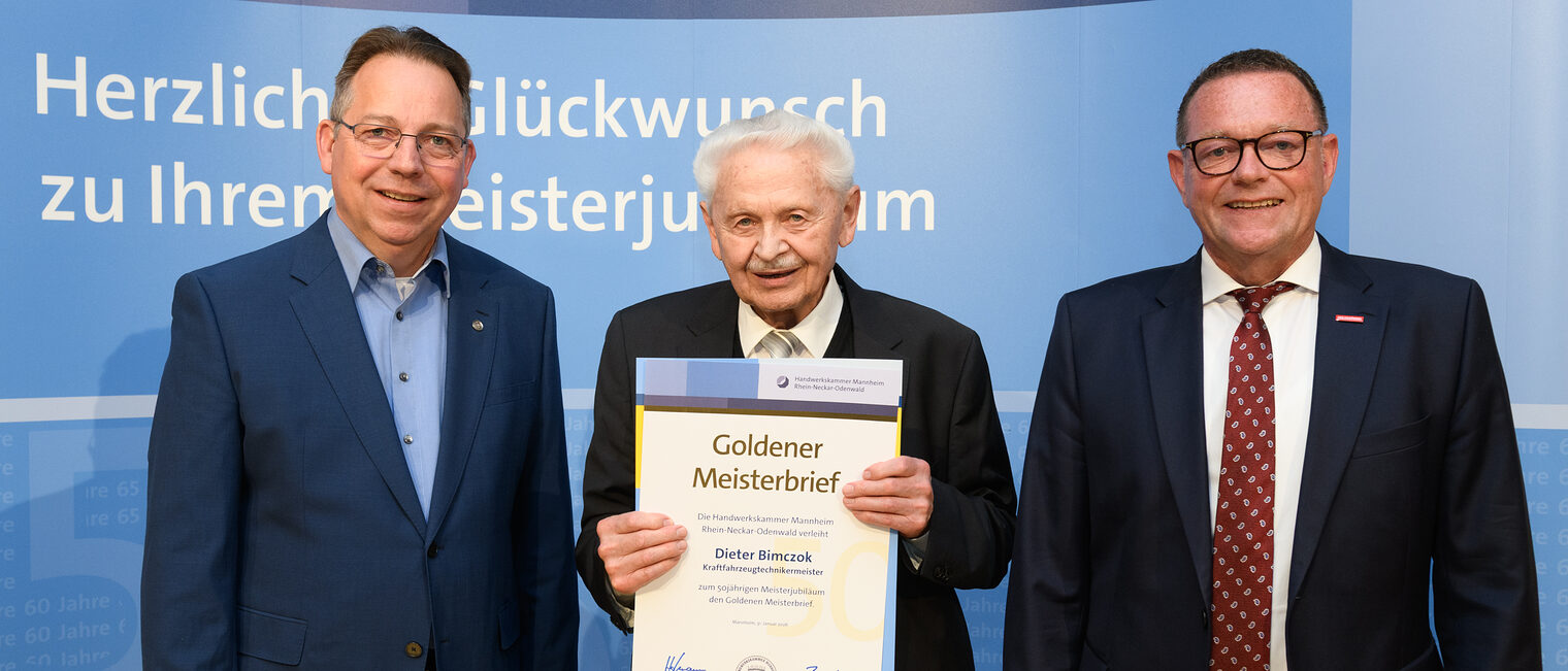 Drei Personen posieren für ein Foto, in der Mitte wird die Ehrenurkunde gehalten