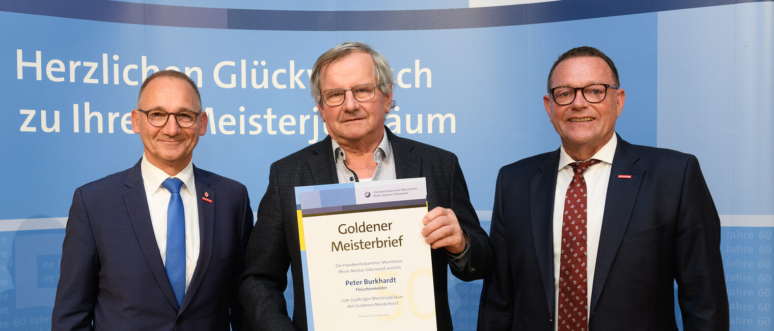 Drei Personen posieren für ein Foto, in der Mitte wird die Ehrenurkunde gehalten