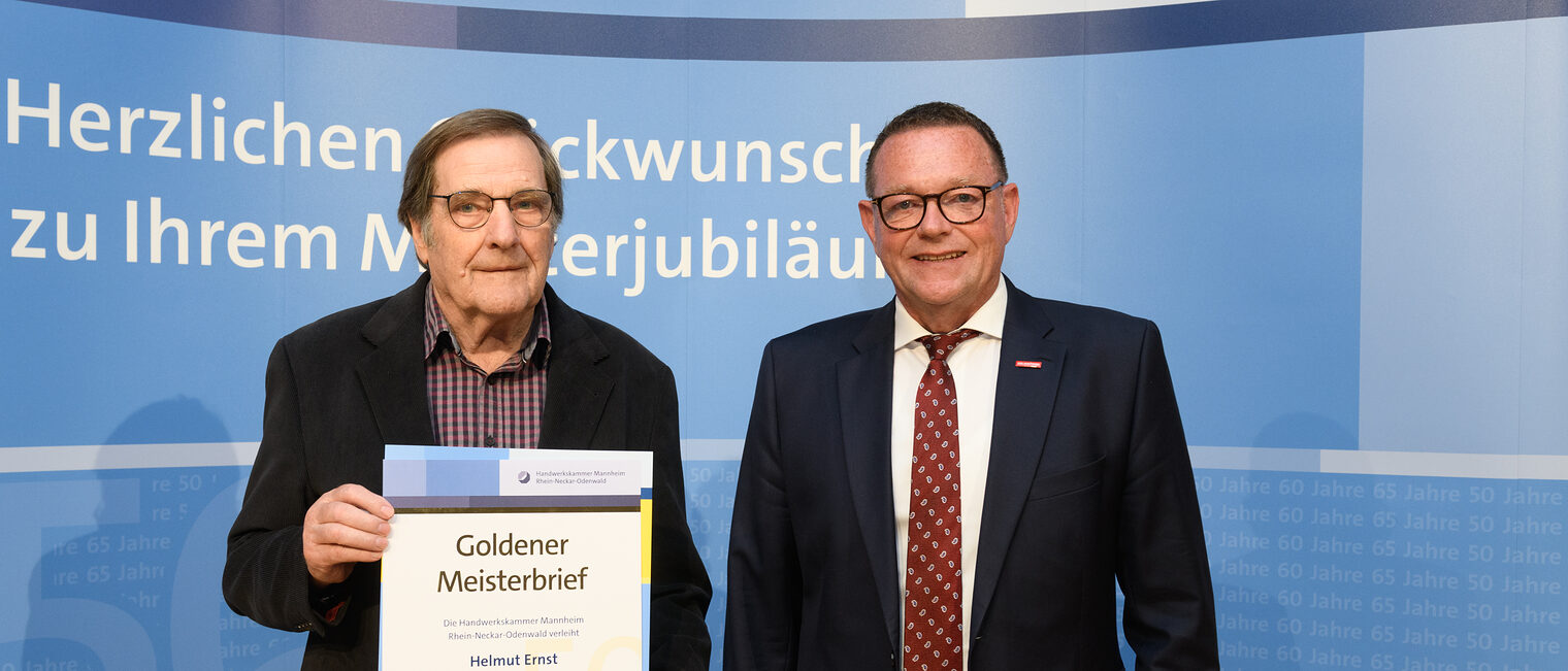 Zwei Personen posieren für ein Foto, in der Mitte wird die Ehrenurkunde gehalten