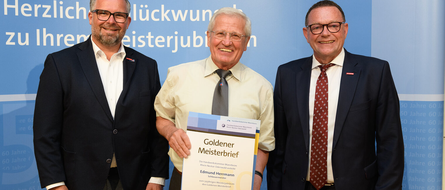 Drei Personen posieren für ein Foto, in der Mitte wird die Ehrenurkunde gehalten