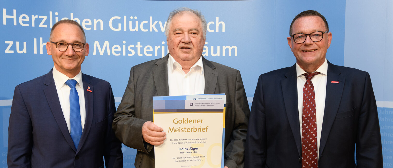 Drei Personen posieren für ein Foto, in der Mitte wird die Ehrenurkunde gehalten