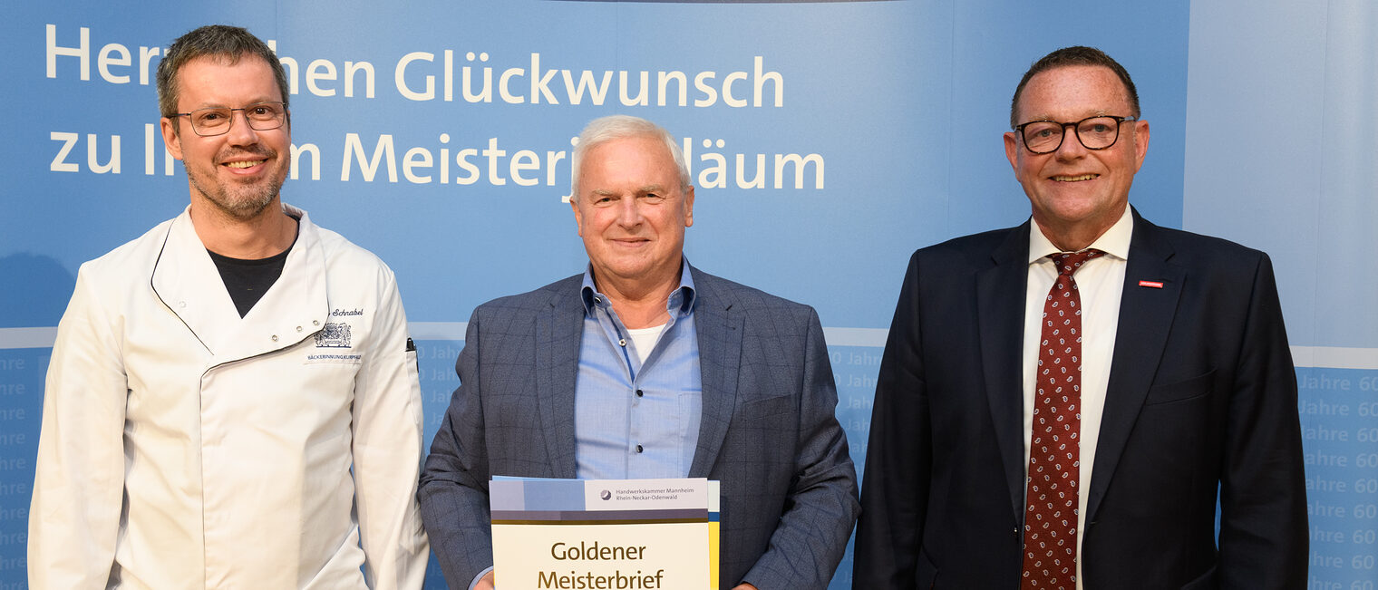 Drei Personen posieren für ein Foto, in der Mitte wird die Ehrenurkunde gehalten