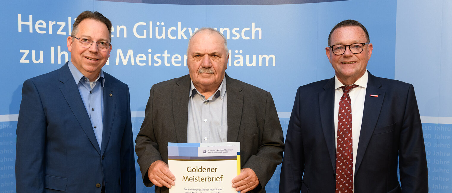 Drei Personen posieren für ein Foto, in der Mitte wird die Ehrenurkunde gehalten