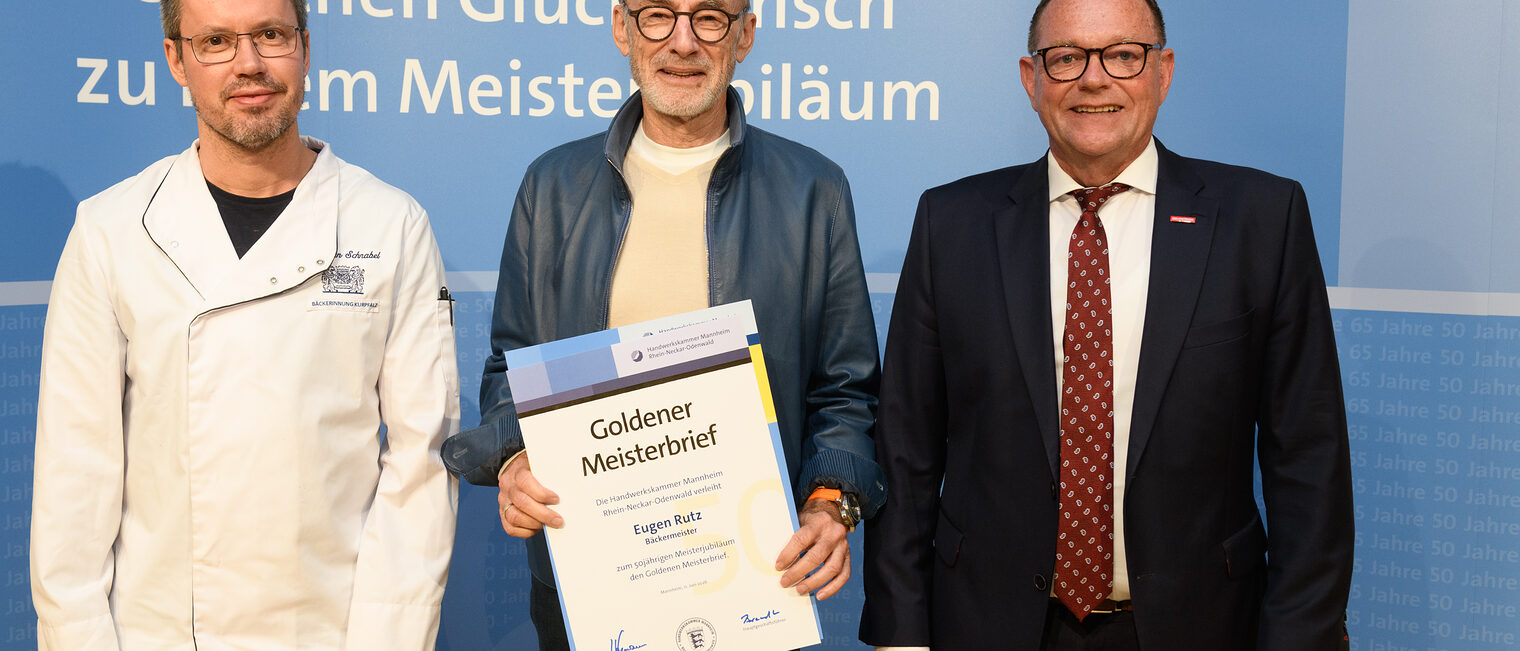 Drei Personen posieren für ein Foto, in der Mitte wird die Ehrenurkunde gehalten