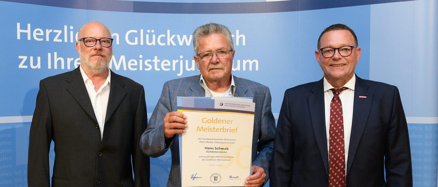 Drei Personen posieren für ein Foto, in der Mitte wird die Ehrenurkunde gehalten